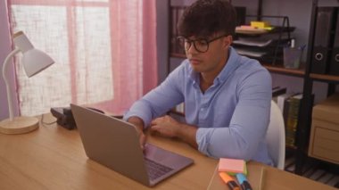 Ofiste çalışan genç İspanyol bir adam pembe perdeler, lamba ve ofis malzemeleriyle çevrili bir dizüstü bilgisayarla oturuyor ve hayal kırıklığını gösteriyor..