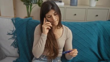 Genç, çekici İspanyol bir kadın, evindeki rahat oturma odasında kredi kartına bakarken telefonda konuşuyor..