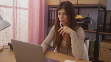 Evinden ofis ortamında çalışan genç bir esmer kadın. Masasında dizüstü bilgisayarla otururken düşünceli görünüyor. Etrafı dosyalar, raflar ve ofis malzemeleriyle çevrili..