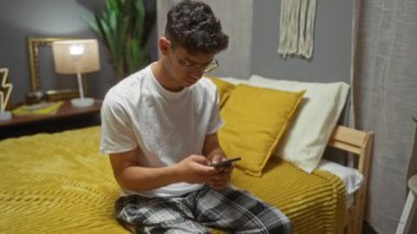 Latin kökenli genç bir adam rahat bir yatak odasında yatağında oturuyor, telefonuna odaklanmış, arkasında sarı bir yatak örtüsü ve komodiniyle..