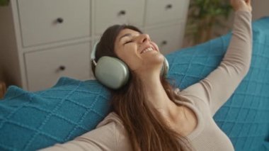 Genç bir esmer kadın kulaklık takıyor ve mavi dekorlu rahat bir oturma odasında gülümsüyor..