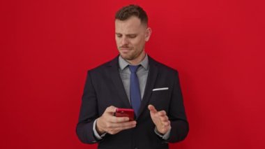 Takım elbiseli genç adam akıllı telefonundaki bir sorundan dolayı somurtuyor, izole edilmiş, kırmızı arka planda negatif duyguları canlandırıyor.