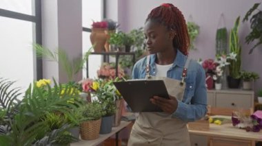 Genç bir Afro-Amerikalı kadın canlı bir kapalı çiçek dükkanında bir pano dikkatle okuyor, yemyeşil ve çiçeklerle çevrili..