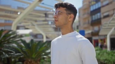 Güneşli bir şehir parkında, arka planda palmiye ağaçları ve modern binaları olan beyaz gömlek giyen, gözlüklü yakışıklı İspanyol bir adam..