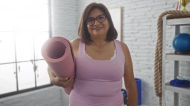 Kapalı spor salonunda kısa saçlı, yoga minderi tutan, gözlük takan ve kameraya gülümseyen yaşlı İspanyol bir kadın..