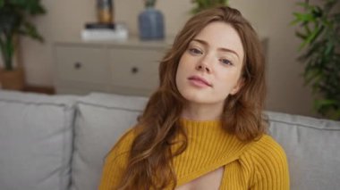 Sarı saçlı, gözlüklü güzel bir kadın evdeki rahat bir oturma odasında oturuyor, gözlüğünü tutarken poz veriyor ve gülümsüyor..