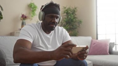 Kulaklık takan Afrikalı genç bir Amerikalı modern bir oturma odasında oturmuş endişeli bir ifadeyle akıllı telefonuna odaklanmış..