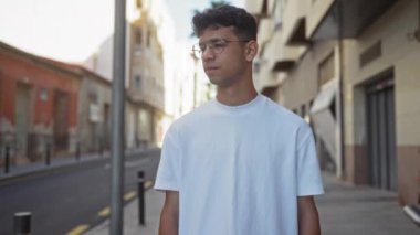Gözlüklü ve beyaz tişörtlü İspanyol genç adam. Arka planda binalar ve yollar olan bir şehir sokağında güven içinde poz veriyor..