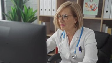 Yetişkin bir kadın doktor, masasında otururken kollarını kaldırarak kutluyor. Parlak, modern bir klinikte, steteskopu boynunda ve rafları arka planda.. 