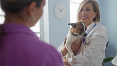 Bir başka kadın ön planda durup evcil hayvan sağlığını önemserken, klinik odasında küçük bir köpeği tutan olgun sarışın bir veteriner..