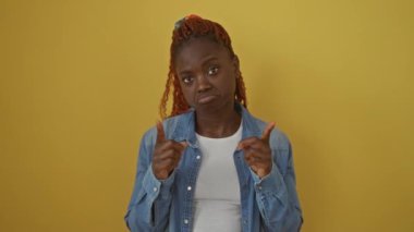 Depresyondaki Afro-Amerikan kadın kot gömlek giyiyor, izole edilmiş sarı bir arkaplan üzerinde dikiliyor ve yukarı doğru işaret ediyor, üzgün ve mutsuz görünüyor üzgün bir ifadeyle