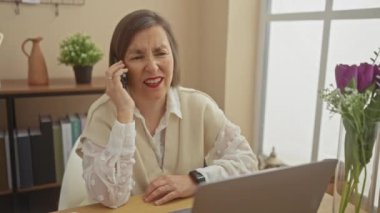 Çağdaş ev ofis ortamında çiçeklerle bilgisayarla çalışan olgun bir kadın telefonda konuşuyor.
