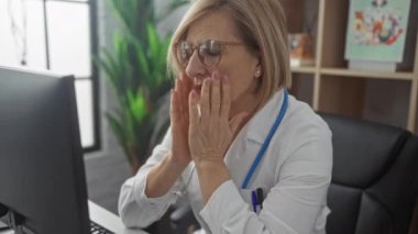Gözlüklü, sarışın, beyaz önlüklü olgun bir kadın doktor modern bir klinikte masa başında otururken yüzünü elinde tutuyor..