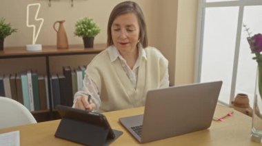 Olgun bir kadın dizüstü bilgisayarında, iyi aydınlatılmış bir ofiste tablet, gözlük ve dekoratif bitkilerle çalışıyor..