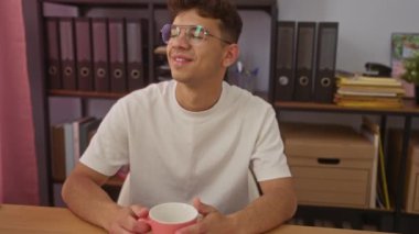 Ofis ortamındaki genç bir İspanyol, elinde pembe bir kupayla iş yerinde düşünceli bir şekilde gülümsüyor..