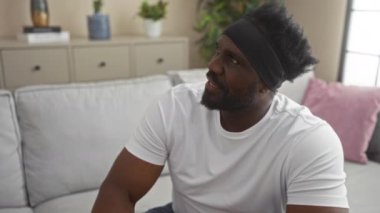 Yakışıklı, sakallı Afrikalı Amerikalı bir adam şık bir oturma odasında, arka planda bitki ve dekor olan bir koltukta oturuyor..