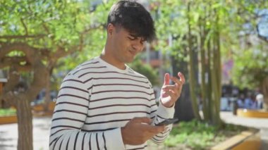 Şehir parkında çizgili tişörtlü genç İspanyol bir adam endişeli bir ifadeyle akıllı telefonuna bakıyor..