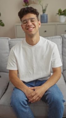Beyaz tişörtlü ve kot pantolonlu gülümseyen genç bir adam, kapalı bir koltukta oturuyor, arka planda bitkiler ve dekorla rahat bir oturma odasında kollarını kavuşturuyor..