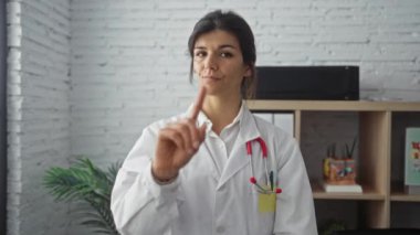 Bir klinikte, esmer bir kadın doktor parmaklarıyla hayır işareti yapar..