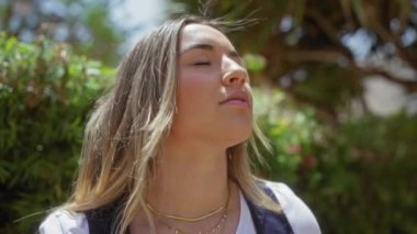 Huzurlu genç bir kadın, güneşli bir şehir parkında güneşli havanın tadını çıkarır, sarı saçları rüzgarda dalgalanır ve gözleri neşeyle kaplanır..