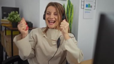 Genç, çekici, sarışın bir kadın iş yerinde kulaklık takarak ofis ortamında çalışırken sevinçle kutlama yapıyor..