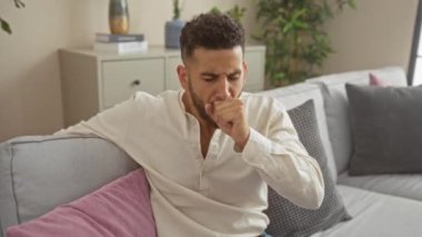 Genç bir İspanyol adam oturma odasında bir kanepede otururken öksürüyor, arka planda çeşitli bitkiler ve dekorlarla evdeki hastalık sahnesini canlandırıyor..