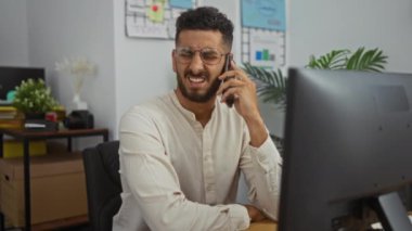 İspanyol, genç, ofis ortamında telefonla konuşan, yakışıklı, yetişkin bir adam bilgisayarlı, çeşitli bitkilerle, iş yeriyle, kapalı bir masada oturuyor..