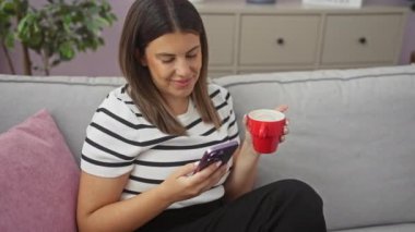 Genç, çekici İspanyol bir kadın rahat oturma odasında oturmuş kırmızı bardaktan kahve yudumluyor ve pembe ve gri yastıklı bir kanepede otururken telefonuna bakıyor..