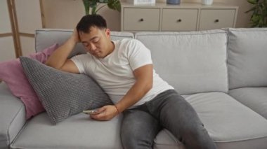 Genç Çinli bir adam rahat bir oturma odasında kanepede uzanıyor, telefonunu kullanıyor ve uykulu görünüyor..