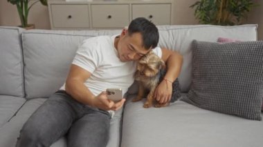 Beyaz tişörtlü genç bir Asyalı oturma odasındaki gri koltukta oturuyor, akıllı telefonunu evde köpeğine sarılırken kullanıyor..