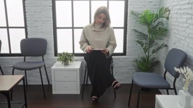 Kapalı bir bekleme odasında tek başına oturan genç sarışın bir kadın modern mobilyalarla dijital tabletle meşgul..