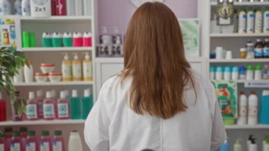 Orta yaşlı gözlüklü ve laboratuvar önlüklü bir kadın bir eczanede duruyor ve arka planda bir sürü farklı ürün var..
