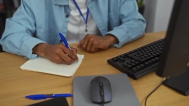 Ofis sahnesinde, bilgisayarın yanındaki not defterine notlar yazan Afro-Amerikan bir adam profesyonel bir iş ortamını gözler önüne seriyor..