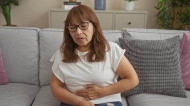 Oturma odasında oturan yaşlı bir İspanyol kadın karın ağrısı çekiyor. İyi aydınlatılmış bir evde, bir kanepede otururken karnını sıkıca tutuyor..