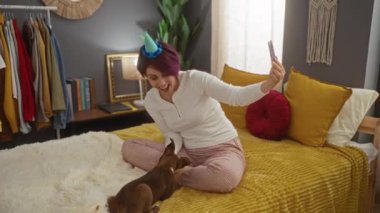 Parti şapkalı genç bir kadın köpeğiyle güzel dekore edilmiş yatak odasında rahat bir yatakta selfie çekerken gülümsüyor..