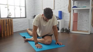 Genç İspanyol bir adam spor salonunda mavi bir paspasın üzerine uzanıyor ve kapalı bir spor merkezinde egzersiz yapıyor..