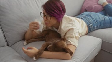 Mor saçlı genç bir kadın oturma odasındaki gri bir kanepede uzanmış, burnuna mendil tutarken köpeğine sarılıyor..