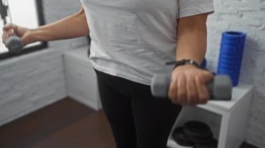 Yaşlı bir kadın spor salonunda elinde ağırlıklarla egzersiz yapıyor, etrafını spor aletleri sarmışken odaklanmış bir ifade sergiliyor..