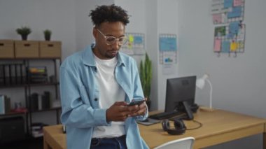 Yakışıklı, Afro-Amerikan bir adam ofis işyerinde duruyor, akıllı telefonunu kullanıyor, odaklanmış bir ifadeyle, modern ofis mobilyası ve dekorasyonuyla çevrili..