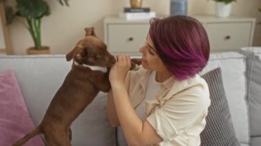 Mor saçlı genç bir kadın, rahat bir oturma odasında kahverengi bir köpekle oynuyor..