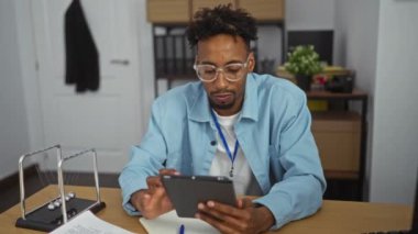 Genç, yakışıklı, sakallı Afrikalı Amerikalı bir adam ofis ortamında bir tablet üzerinde çalışıyor. Etrafı evrak ve masa aksesuarlarıyla çevrili..