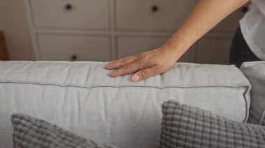 Yaşlı bir İspanyol kadın bir evin içinde elini nazikçe gri bir kanepeye koyar. Gri yastıklar ve bulanık bir oturma odası dolabının arka planı vardır..