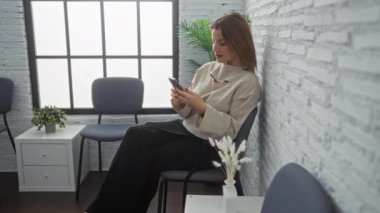 Modern bir bekleme odasında minimalist dekorla otururken akıllı telefon kullanan çekici genç bir kadın, güneşli bir günde bitkiler ve mavi sandalyeler..