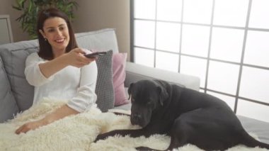 Latin kökenli genç bir kadın, siyah labrador köpeğiyle bir kanepede dinleniyor, rahat, modern bir evde televizyon seyrediyor..