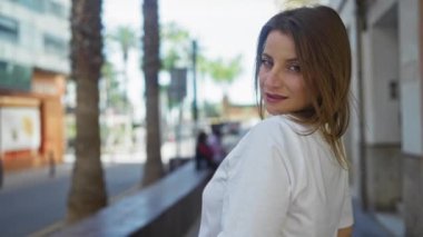 Güneşli bir günde arka planda binalar ve palmiye ağaçları olan bir caddede gülümseyen genç, çekici bir kadın..