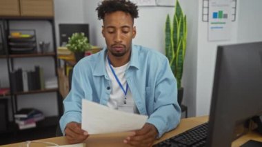 Ofiste sakallı, elinde belgelerle çalışan mavi bir iple arka planda bilgisayar ve ofis malzemeleriyle bir masada oturan Afrikalı genç bir Amerikalı..