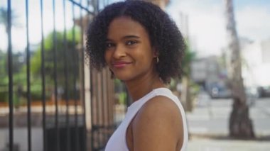 Kıvırcık saçlı, güzel bir Afro-Amerikalı kadın, güneşli bir günde şehir çevresi ile çevrili beyaz bir atlet giymiş, bir sokak ortasında gülümsüyor..