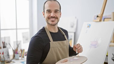 Smiling bearded man painting on canvas in bright art studio wearing apron clipart