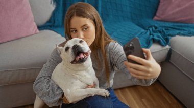 Genç ve güzel sarışın bir kadın, beyaz Fransız bulldoguyla sıcak bir oturma odasında selfie çekiyor..