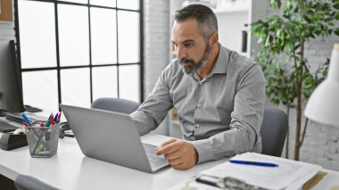 Modern ofis ortamında dizüstü bilgisayarla çalışan gri sakallı olgun bir İspanyol.